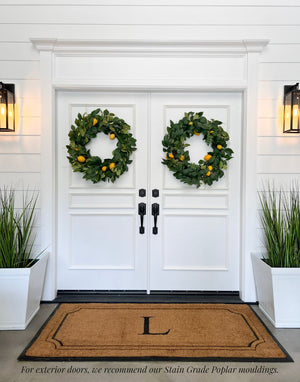 White double entry doors featuring the three piece door moulding kit, showcasing a classic paneled design framed by white trim and a door pediment, styled with green wreaths and exterior wall lighting.